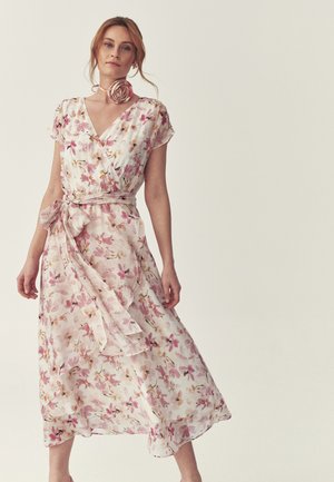 Frau trägt ein hellrosa geblümtes Wickelkleid mit gebundenem Taillenband und einem passenden geblümten Choker, steht vor einem schlichten Hintergrund.