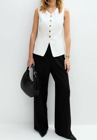 Bílý upravený vest s zlatými knoflíky, v kombinaci s černými kalhotami širokého střihu a černou kabelkou. Hladký, moderní design a ostré linie.
