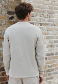 Vaaleabeige pitkähiha-paita, jossa on teksturoitu vohvelikuvio, pyöreä pääntie ja hieman kaartuva helma, yhdistetty vaaleisiin housuihin.