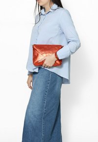Sac à main en métal rouge avec une surface texturée, doté d'une fermeture éclair et d'un design angulaire. Assortie avec une chemise bleu clair et une jupe en jean.