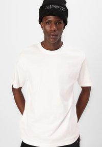 Weißes Baumwoll-T-Shirt mit kurzen Ärmeln und rundem Ausschnitt, getragen mit einer schwarzen Strickmütze mit weißer Schrift. Schlichtes Design, keine Muster.