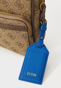 Geantă de umăr cu model maro, dotată cu un buzunar frontal cu fermoar și o etichetă din piele albastră atașată, inscripționată cu "GUESS" în litere aurii.