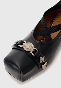 Black leather shoe with a square toe design, featuring silver hardware accents and crossed black elastic straps. Smooth texture.