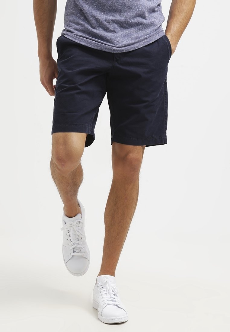 Muž kráčející v tmavě modrých šortkách, šedém tričku a bílých teniskách, s jednou rukou v kapse na bílém pozadí.