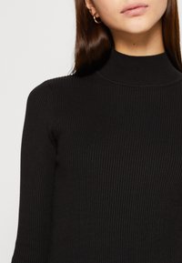 Musta ribbineulottu poolopaita pitkähihaisilla hihoilla, jossa on istuva malli ja sileä pinta, täydentäen yksinkertaiset kultaiset renkaankorvakorut.