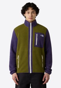 Giacca in pile di colore verde oliva e viola, con colletto alto, tasche con zip e un logo sulla tasca del petto. Tessuto testurizzato.