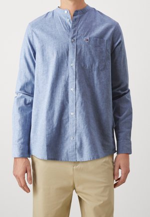 Hombre con camisa azul claro de cuello mandarín con botones y bolsillo en el pecho, combinada con pantalones beige.