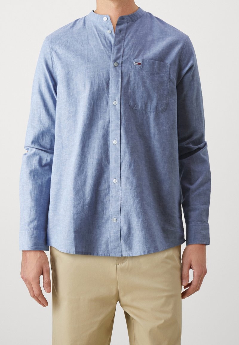 Homme portant une chemise bleu clair à col mandarin avec des boutons et une poche poitrine, associée à un pantalon beige.