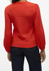 Rød tekstureret langærmet top med en scoop neckline, der har samlede ærmer og en tætsiddende talje, parret med mørkeblå jeans.