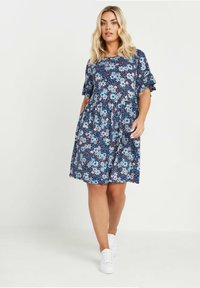 Marinblommig klänning med korta volangärmar, prydd med blå och vita blommor på en prickig bakgrund. Bärs med vita sneakers.