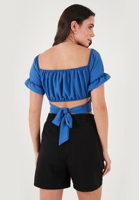 Blå cropped top med puffærmer, elastisk talje og sløjfedetalje på ryggen. Kombineret med sorte shorts, der fremviser et tætsiddende design.
