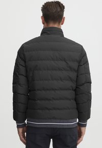 Svart pufferjakke med quilted design, høy krage og elastiske mansjetter. Har en ribbestrikket midjebånd med grå og hvite striper.