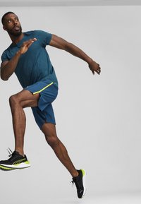 Modelo masculino atlético con una camiseta de manga corta turquesa y pantalones cortos azules con detalles amarillos, corriendo en zapatillas negras y verde neón.