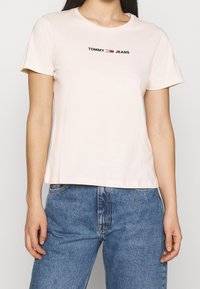 Camiseta de algodón de color rosa claro con cuello redondo, mangas cortas y el logo "TOMMY JEANS" en negro y rojo en el pecho. Corte estándar.