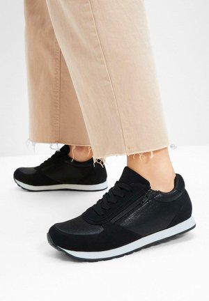 Baskets noires avec un mélange de textures mates et brillantes, semelle blanche, design à lacets et zips sur les côtés. Portées avec un pantalon clair effiloché.