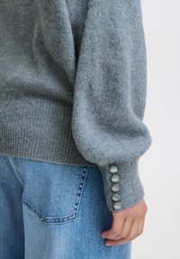 Maglione grigio lavorato a maglia con una finitura testurizzata, caratterizzato da maniche a sbuffo e cinque bottoni di colore chiaro sul polsino. Pantaloni in denim visibili sotto.
