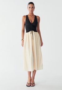 Svart ribbad topp med djup V-ringning och rynkdetaljer, kombinerad med en luftig crèmefärgad kjol med ett strukturerat rutmönster. Svarta flip-flops.