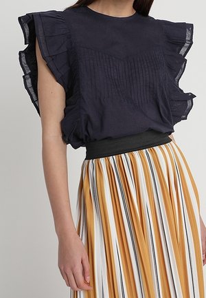 Vrouw draagt een marineblauwe, ruchesloze mouwloze blouse, ingestopt in een geplooide high-waist rok met verticale mosterd-, witte en zwarte strepen.