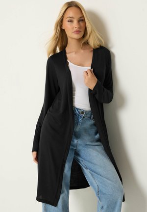 Mujer rubia que lleva una larga rebeca negra abierta sobre una camiseta blanca y unos vaqueros azul claro de cintura alta, sobre un fondo liso y claro.