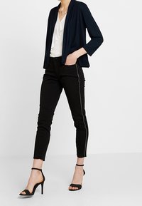 Marinblå 3/4 ärm cardigan över en vit blus, i kombination med svarta skinny jeans med sidosträck och svarta högklackade sandaler.