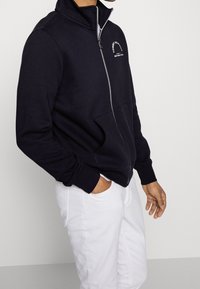 Felpa con zip navy e colletto alto, caratterizzata da un logo bianco e due tasche frontali; indossata con pantaloni bianchi. Materiale in morbido cotone misto.