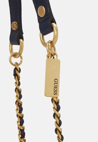 Bracelet en cuir bleu marine avec des accessoires en métal doré, présentant un design de chaîne torsadée et une étiquette rectangulaire portant la mention "GUESS".