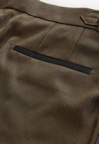 Brązowe spodnie materiałowe z teksturowanym wzorem w diamenty, z pojedynczą czarną kieszenią jako akcentem. Gładkie wykończenie i dopasowany krój.
