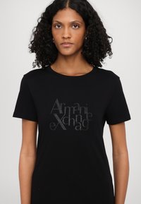 Čierne bavlnené tričko s krátkymi rukávmi a okrúhlym výstrihom, ktoré má lesklé, štýlizované logo "Armani Exchange" v sivej farbe.
