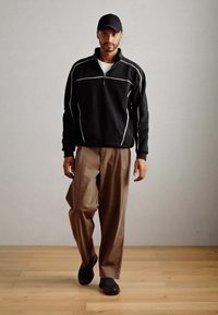 Man walking indoors wearing a black cap, black quarter-zip long sleeve top with white piping, brown wide-leg pants, and black shoes.