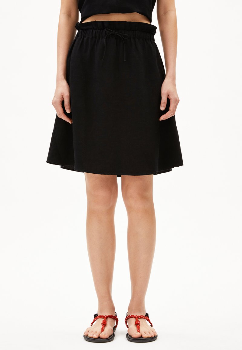 Schwarzer, ausgestellter Rock mit elastischem Bund und Kordelzug. Getragen mit roten Sandalen mit verknoteten Riemen und flachen Sohlen.