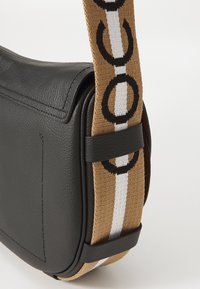 Sort læder messenger taske med en struktureret overflade, der har en bred, mønstret rem i brunt og hvidt med cirkulære designs.