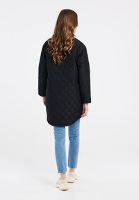 Cappotto lungo trapuntato nero con orlo arrotondato e maniche lunghe, abbinato a jeans azzurri e sneakers beige.