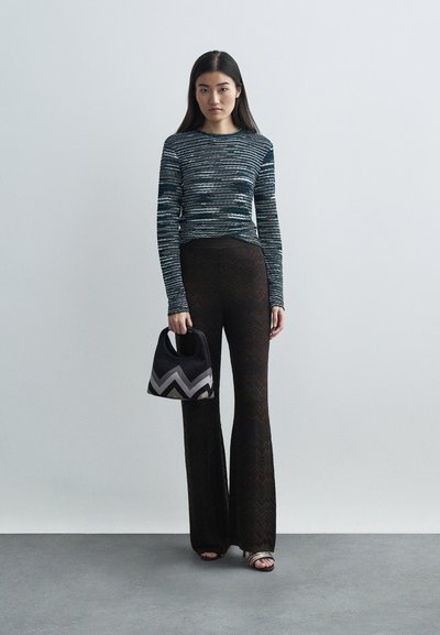 Γυναίκα που φοράει μπλε μοτίβο μπλουζάκι με μακριά μανίκια, μαύρο παντελόνι με ζιγκ-ζαγκ μοτίβο, κρατώντας μια τσάντα ζιγκ-ζαγκ μαύρης και γκρι απόχρωσης, στέκεται σε γκρίζο πάτωμα.