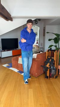 Jeune homme en sweat à capuche bleu et jean, debout dans un salon avec un canapé marron, une télévision, une plante et un rameur en bois.
