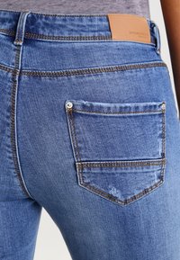 Vyšisované modré džíny vyrobené z denimu, s páskem střední výšky, oranžovým prošíváním a malou zadní kapsou s kovovým nýtem. Náprsní nášivka na pásku.