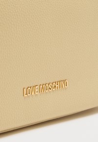 Μπεζ τσάντα από ψεύτικο δέρμα με υφή, που φέρει το εντυπωσιακό χρυσαφί λογότυπο "LOVE MOSCHINO" στο μπροστινό μέρος. Ομαλή επιφάνεια, μίνιμαλ σχεδιασμός.
