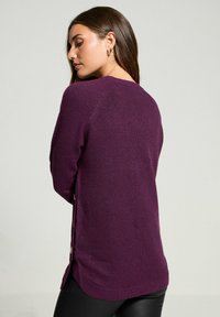 Lila Pullover mit entspannter Passform, gerippter Textur, seitlichen Knopfakzenten und leicht abgerundetem Saum. Das Material wirkt weich und gestrickt.