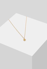Zelta ķēdīte ar mazu, apaļu piekariņu ar teksturētu dizainu, kas izlikts minimālistiskā pelēkā fonā.