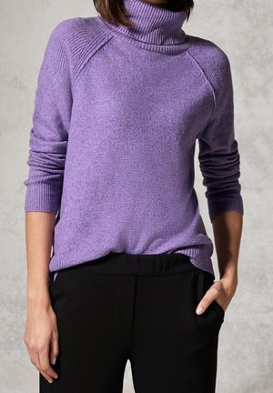 Pull à col roulé violet avec col et poignets côtelés, porté avec un pantalon noir, une main dans la poche, sur un fond gris clair.