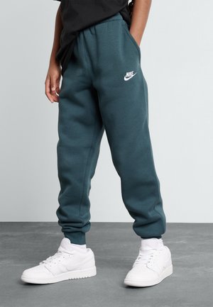 Person iført mørk teal Nike joggingbukser og hvide Nike sneakers, stående på gråt gulv mod en lysegrå baggrund.