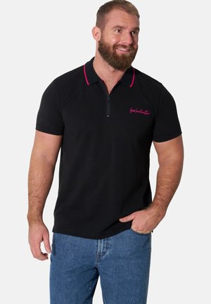 Bearded man wearing black short-sleeve polo shirt with pink piping and blue jeans, smiling and looking to the side with one hand in pocket.