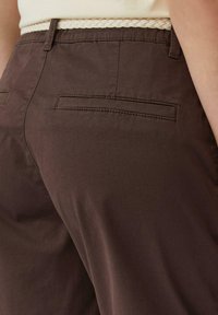 Bruine katoenen broek met twee achterzakken en een gevlochten lichtgekleurde riem. De stof heeft een gladde textuur en een comfortabele pasvorm.