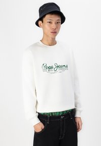 Sudadera blanca con el logo verde de "Pepe Jeans", combinada con pantalones negros y una cinturilla de cuadros verdes. Modelo lleva un sombrero tipo bucket negro.