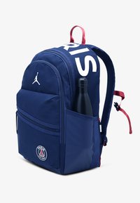 Navy blue backpack with a large white "PARIS" strap, side pocket holding a black water bottle, and Paris Saint-Germain logo on front.