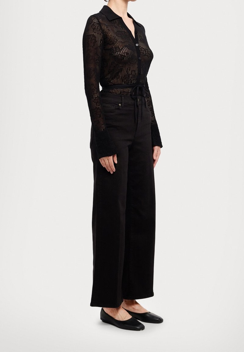 Haut noir à manches longues avec boutons, en tissu transparent à motifs ; assorti à un pantalon noir taille haute, jambes larges et des ballerines noires.