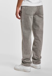 Personne portant un jean gris ample et des baskets blanches, debout contre un fond clair uni, vue de dos et de profil.
