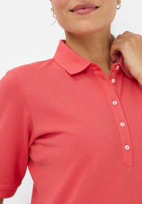 Koralfarvet polo-shirt lavet af struktureret stof med en klassisk krave, som har en tre-knaps placket og kontrasterende hvide knapper.