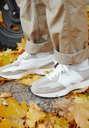 Stopy w białych i beżowych trampkach oraz beżowych spodniach stojące na ziemi pokrytej żółtymi opadłymi liśćmi jesieni.