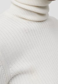 Pull à col roulé côtelé blanc avec un col haut replié et une fine texture verticale. Le tissu semble doux et léger.