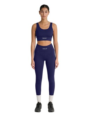 Junge Frau, die nach vorne schaut, trägt einen passenden dunkelblauen Sport-BH und Leggings mit "HALO"-Logo, schwarze Turnschuhe und weiße Socken.